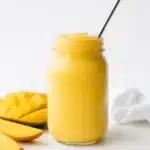 Creamy mango drink in a glass jar with a metal straw on a light background with fresh fruit slices
