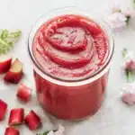 Jar of thick red rhubarb mixture with soft swirl texture on top placed on a light surface