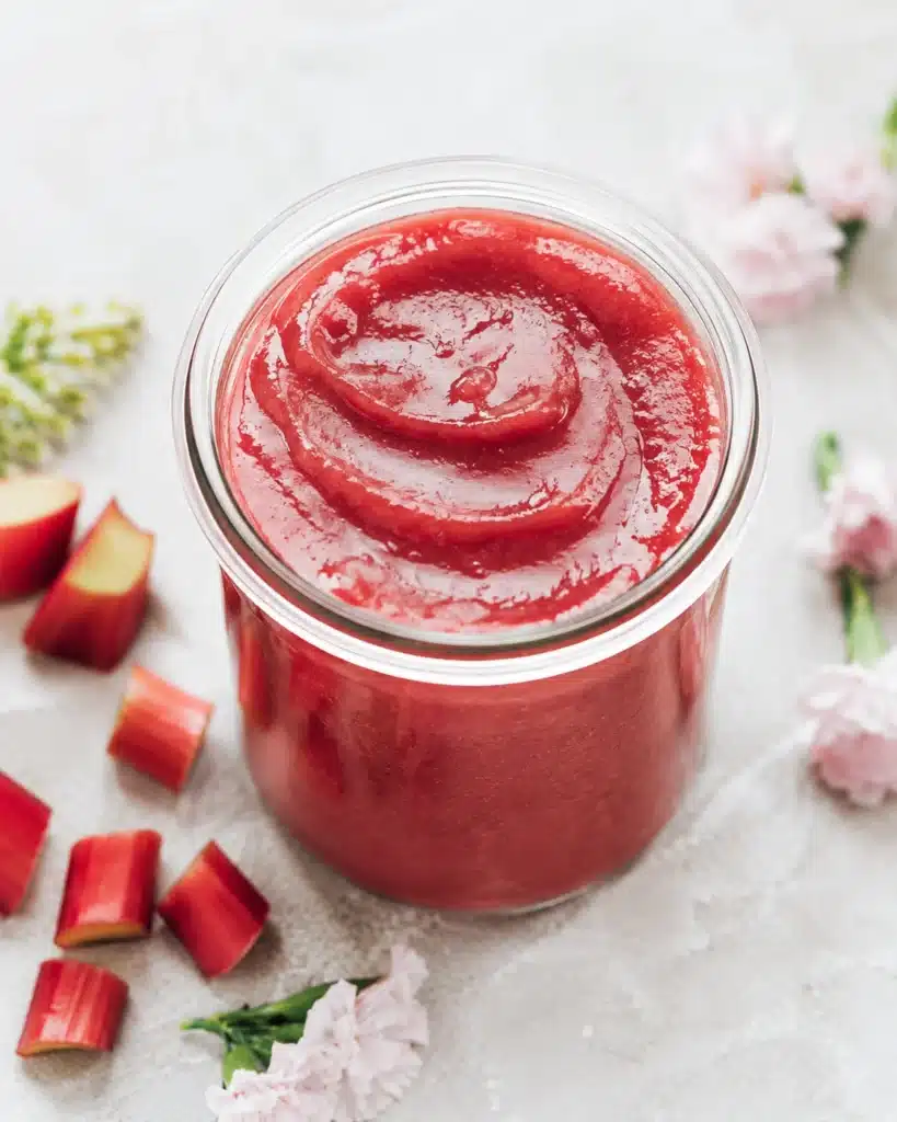 Jar of thick red rhubarb mixture with soft swirl texture on top placed on a light surface