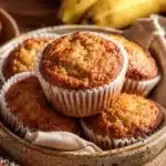 Golden banana muffins in a ceramic bowl with ripe bananas in the background