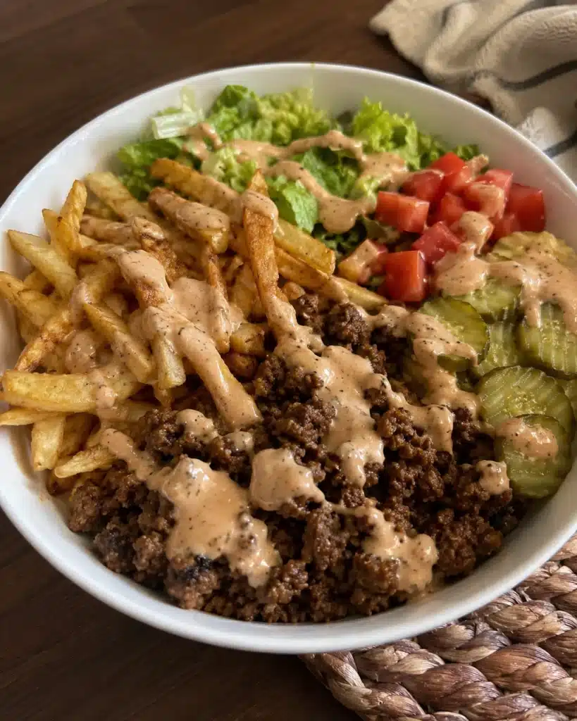 Homemade bowl with ground beef, fries, lettuce, tomatoes, pickles, and creamy sauce on wooden table