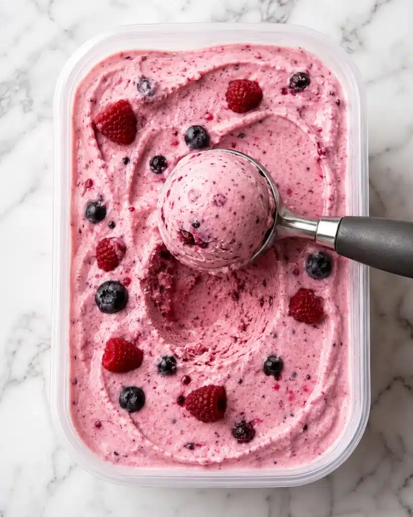 Creamy pink berry yogurt dessert in a container with a scoop and fresh raspberries and blueberries on top
