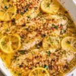Baked tilapia fillets in a white dish with garlic butter sauce, lemon slices, and fresh herbs