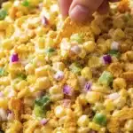hand dipping a corn chip into creamy corn salad with peppers and onion
