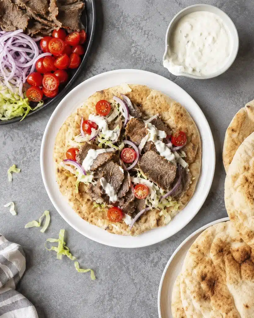 Open pita topped with sliced beef, lettuce, cherry tomatoes, red onion, and creamy white sauce.