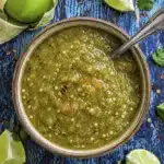 close up of tomatillo enchilada sauce in a bowl with spoon and fresh tomatillos on a rustic surface